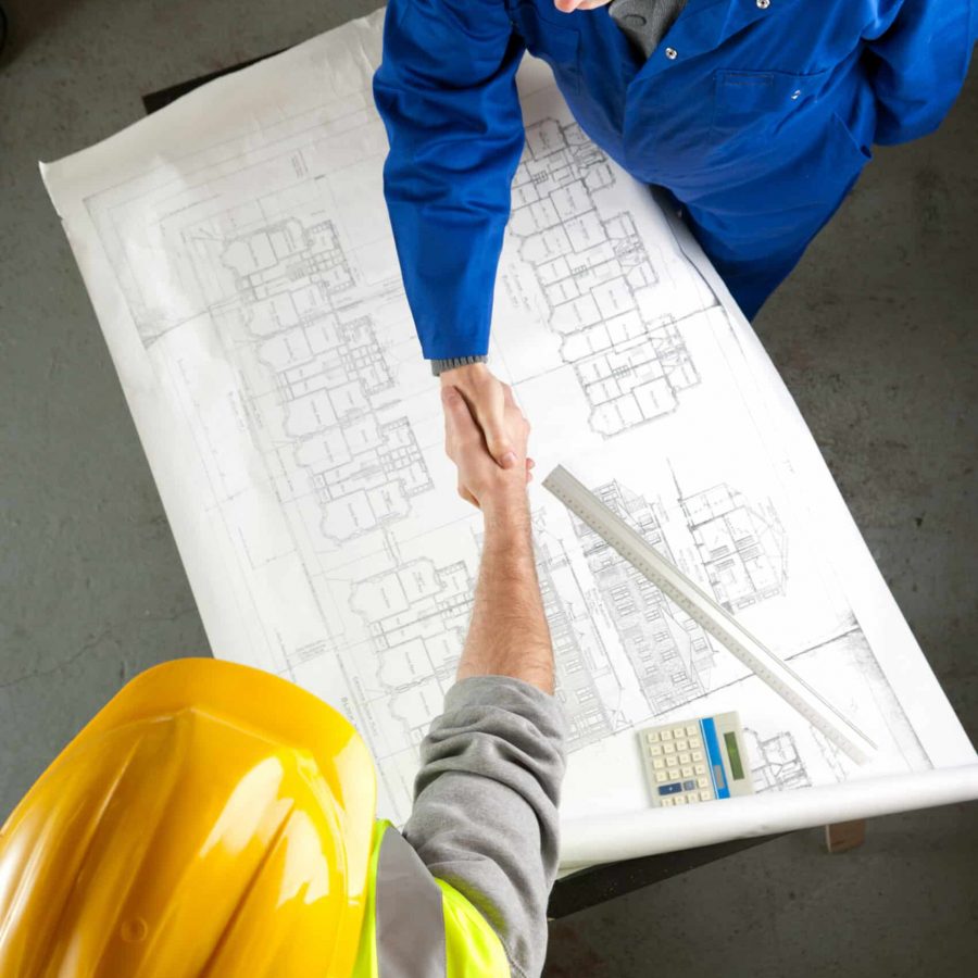Two builders discussing construction plans on a bench in a workshop shake hands on agreement. Overhead shot. Focus on handshake.